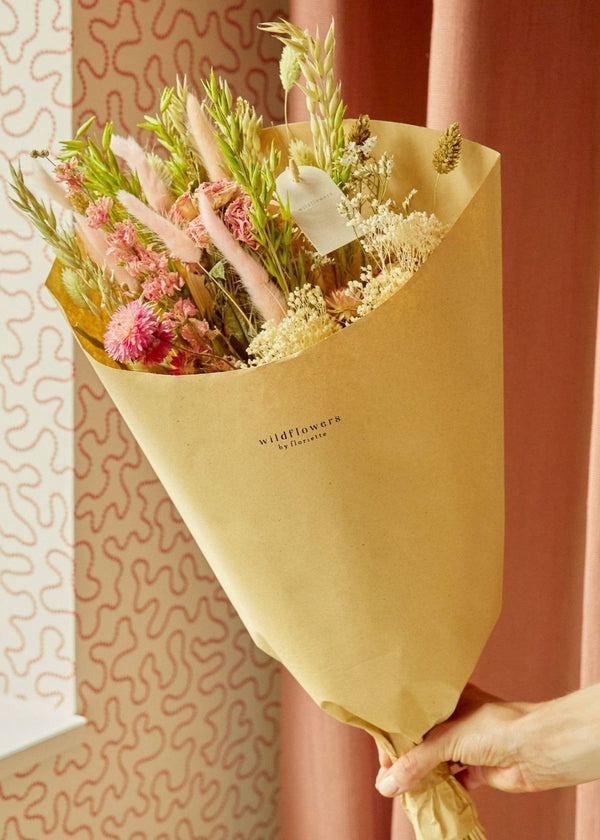 Bouquet of flowers wrapped in yellow paper held by a person against a pink curtain.