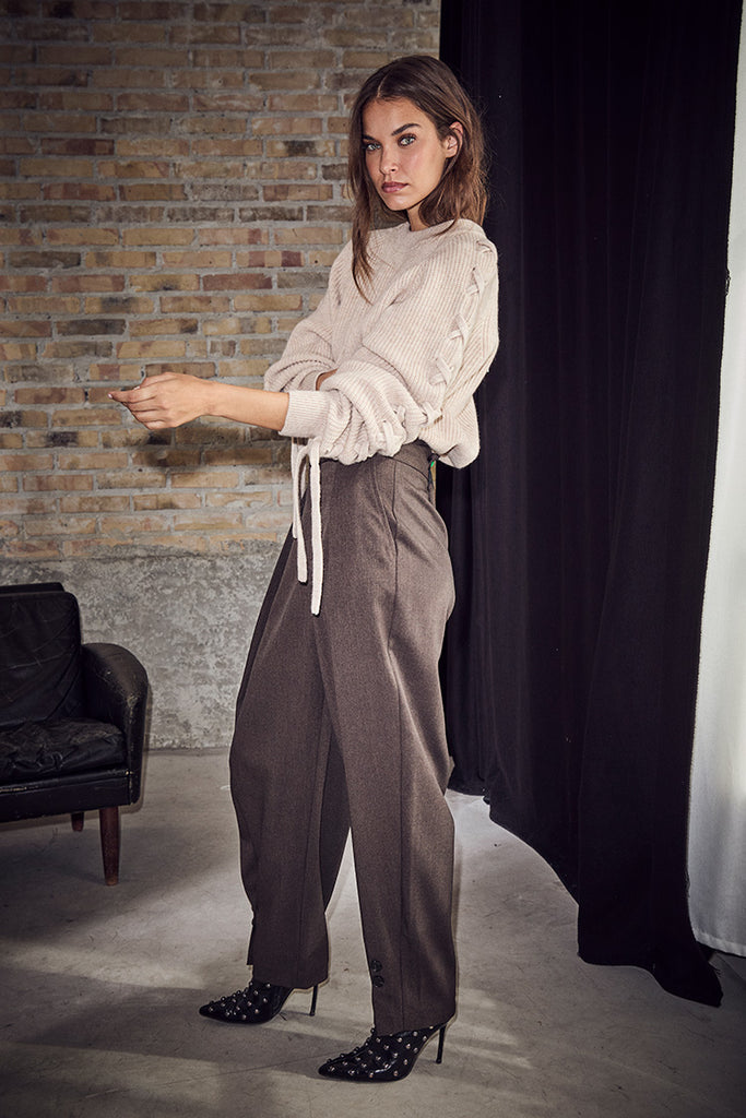 A woman models a two-piece outfit consisting of a cream-colored long-sleeve blouse with ruffled details and high-waisted taupe-brown wide-leg trousers, paired with black polka-dot heels, set against a studio backdrop with a brick wall and black curtain.